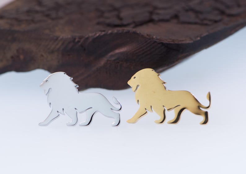 May include: Two lion-shaped metal pins. One is silver-colored, the other is gold-colored. Both pins are side-profile depictions of a lion with a flowing mane and a curled tail. The pins are on a white surface with a dark brown, textured background.