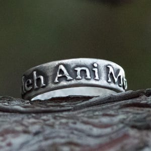 Puede incluir: Primer plano de un anillo de plata con la inscripci&oacute;n "leh Ani M". El anillo se encuentra sobre un fondo natural borroso. La superficie del anillo tiene un aspecto ligeramente envejecido, con las letras resaltadas.