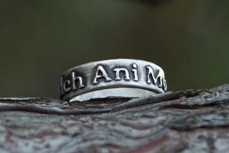 Puede incluir: Primer plano de un anillo de plata con la inscripci&oacute;n "leh Ani M". El anillo se encuentra sobre un fondo natural borroso. La superficie del anillo tiene un aspecto ligeramente envejecido, con las letras resaltadas.