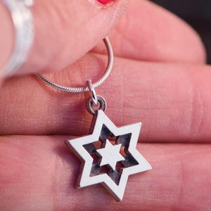 May include: A silver pendant necklace with a Star of David design. The pendant has a layered star shape with a smaller star inside. The necklace is held by a hand with red nail varnish.