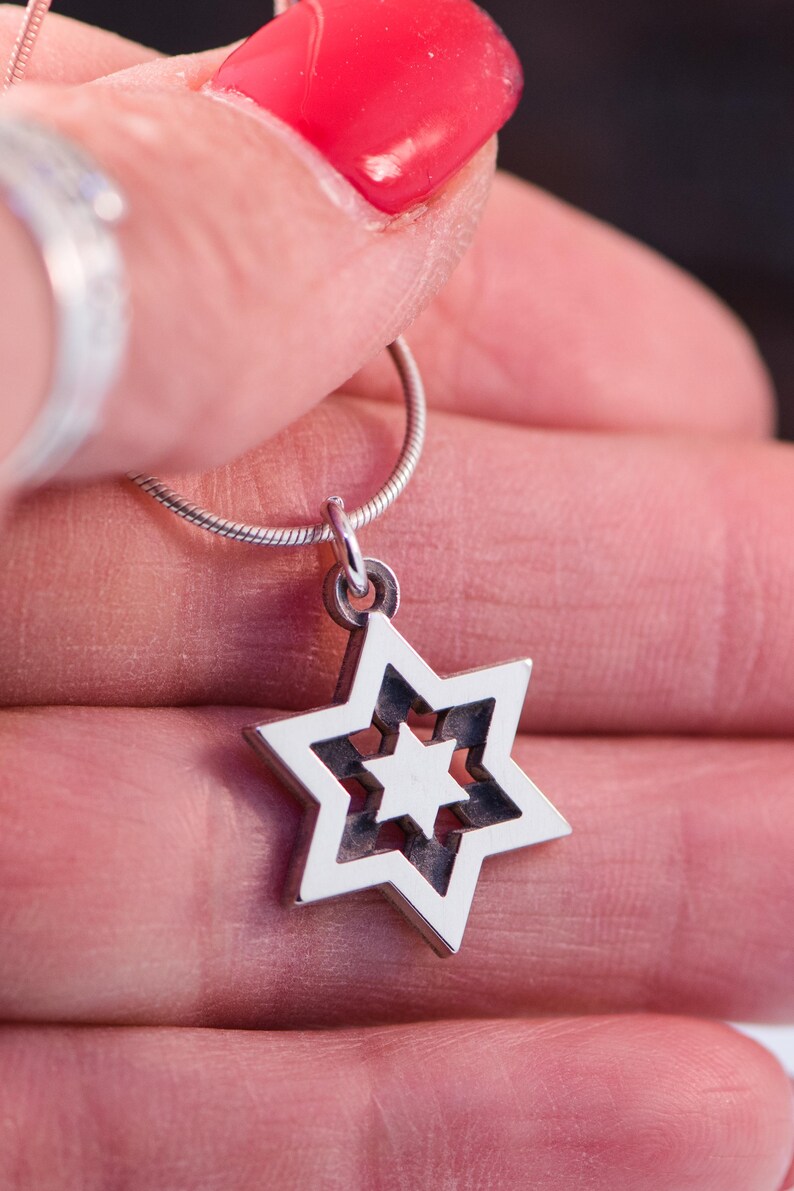 May include: A silver pendant necklace with a Star of David design. The pendant has a layered star shape with a smaller star inside. The necklace is held by a hand with red nail varnish.