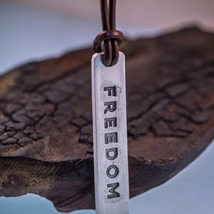 May include: A silver pendant necklace with the word "FREEDOM" engraved vertically. The pendant hangs from a dark brown leather cord. The background is a piece of dark, textured wood.