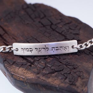 Op de afbeelding: Een zilveren armband met een rechthoekig middelpunt en schakels. Het middelpunt heeft gegraveerde tekst. De armband wordt gepresenteerd op een stuk donker, getextureerd hout.