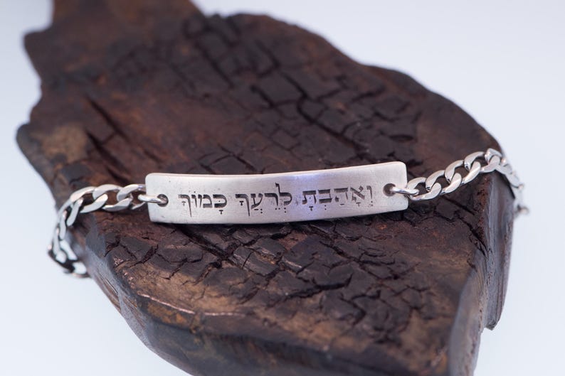 Op de afbeelding: Een zilveren armband met een rechthoekig middelpunt en schakels. Het middelpunt heeft gegraveerde tekst. De armband wordt gepresenteerd op een stuk donker, getextureerd hout.