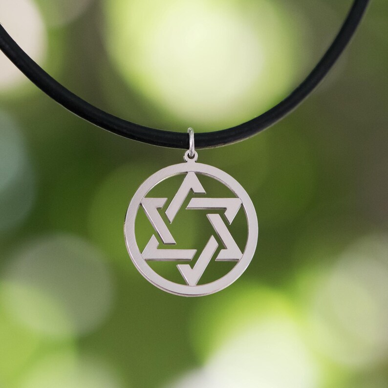 Pu&ograve; includere: Collana con pendente a forma di Stella di David in argento. Il pendente, una stella a sei punte in un cerchio, &egrave; appeso a un cordino nero. Lo sfondo &egrave; verde sfocato.
