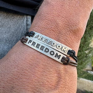 May include: Two silver bracelets with the word "FREEDOM" engraved on them. The bracelets are made of metal and leather, with a dark brown leather band and a silver plate. The bracelets are worn on a wrist.