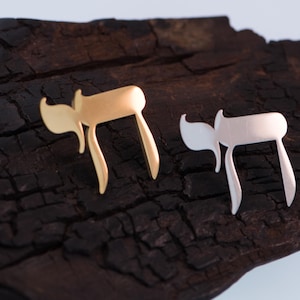 K&ouml;nnte beinhalten: Zwei stilisierte Chai-Anh&auml;nger in Gold- und Silberfarbe. Die Anh&auml;nger werden auf einer dunklen, strukturierten Holzoberfl&auml;che pr&auml;sentiert. Das Chai-Symbol, das im Hebr&auml;ischen "Leben" bedeutet, ist ein beliebtes j&uuml;disches Symbol.
