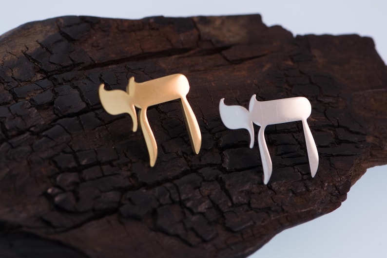 K&ouml;nnte beinhalten: Zwei stilisierte Chai-Anh&auml;nger in Gold- und Silberfarbe. Die Anh&auml;nger werden auf einer dunklen, strukturierten Holzoberfl&auml;che pr&auml;sentiert. Das Chai-Symbol, das im Hebr&auml;ischen "Leben" bedeutet, ist ein beliebtes j&uuml;disches Symbol.