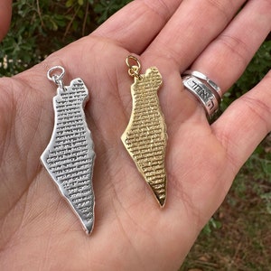 May include: Two pendants in the shape of Israel, one silver-colored and one gold-colored, are held in a hand. Both pendants have detailed text across their surface. A silver ring with Hebrew text is also visible.