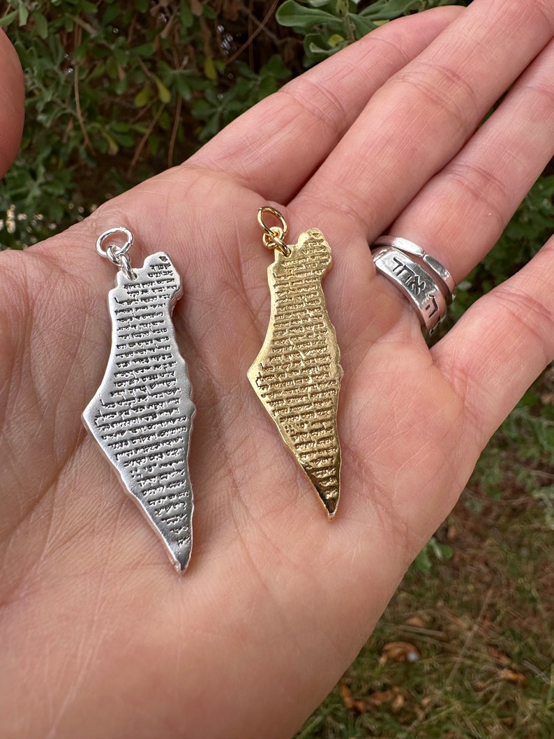 May include: Two pendants in the shape of Israel, one silver-colored and one gold-colored, are held in a hand. Both pendants have detailed text across their surface. A silver ring with Hebrew text is also visible.