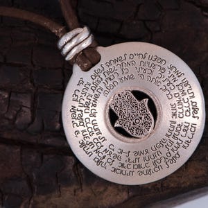 May include: A silver pendant necklace with a circular design and Hebrew text. The pendant features a cutout Hamsa hand symbol in the center and hangs from a brown leather cord.