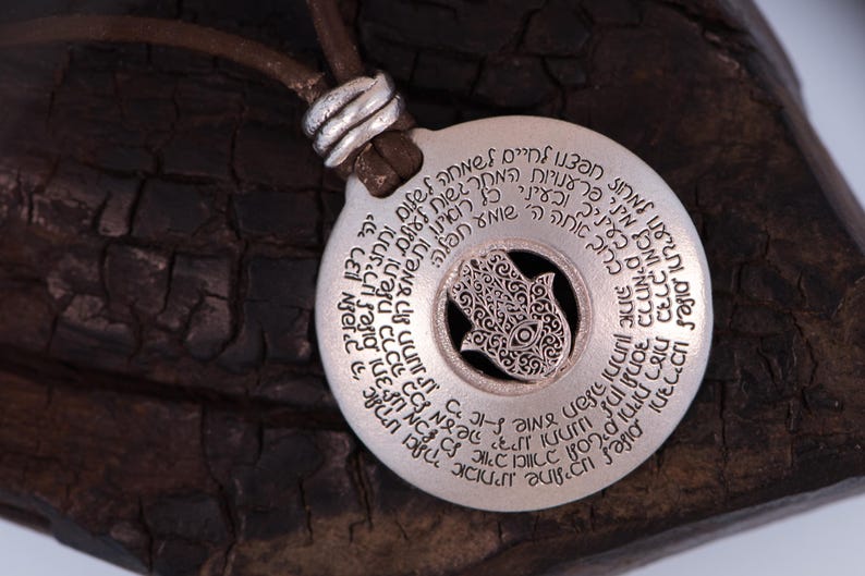 May include: A silver pendant necklace with a circular design and Hebrew text. The pendant features a cutout Hamsa hand symbol in the center and hangs from a brown leather cord.