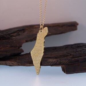 May include: A gold-colored pendant necklace featuring the shape of a country, with text inscribed on the surface. The pendant hangs from a delicate gold chain. The background includes dark, textured wood.