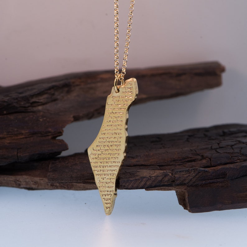 May include: A gold-colored pendant necklace featuring the shape of a country, with text inscribed on the surface. The pendant hangs from a delicate gold chain. The background includes dark, textured wood.