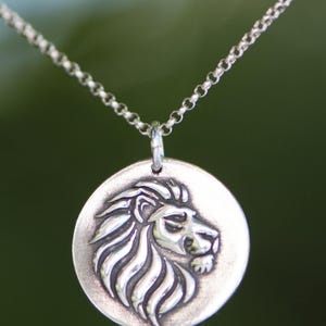 May include: A silver-toned necklace featuring a round pendant with a detailed lion's head in profile. The lion's mane and facial features are intricately carved. The chain is a delicate, linked design, and the background is a blurred green.