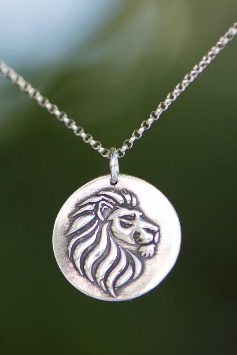 May include: A silver-toned necklace featuring a round pendant with a detailed lion's head in profile. The lion's mane and facial features are intricately carved. The chain is a delicate, linked design, and the background is a blurred green.
