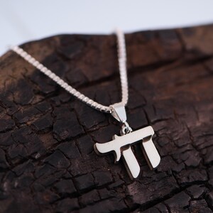 May include: A silver Chai pendant necklace. The pendant is in the shape of the Hebrew letters that spell "Chai", meaning "life". The necklace is displayed on a dark, textured wooden surface.