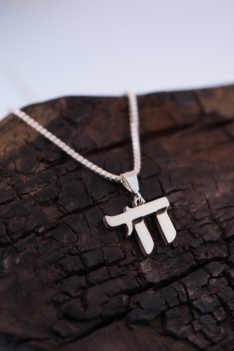 May include: A silver Chai pendant necklace. The pendant is in the shape of the Hebrew letters that spell "Chai", meaning "life". The necklace is displayed on a dark, textured wooden surface.