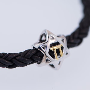 Op de afbeelding: Een gevlochten armband van zwart leer met een zilveren Davidster-bedel met een gouden Hebreeuwse letter 'Chai' erin.