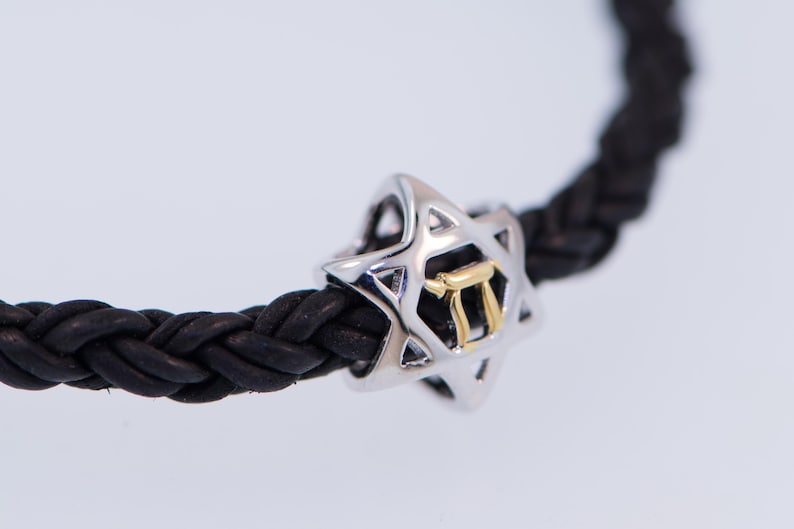 K&ouml;nnte beinhalten: Ein geflochtenes Armband aus schwarzem Leder mit einem silbernen Davidstern-Anh&auml;nger, der einen goldenen hebr&auml;ischen Buchstaben 'Chai' im Inneren tr&auml;gt.