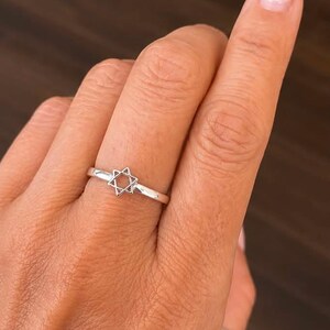 Puede incluir: Un anillo de plata pulida que presenta un peque&ntilde;o s&iacute;mbolo de la Estrella de David calado, centrado en una banda fina y lisa. El metal tiene un acabado met&aacute;lico reflectante y el dise&ntilde;o consta de dos tri&aacute;ngulos entrelazados que forman la estrella.