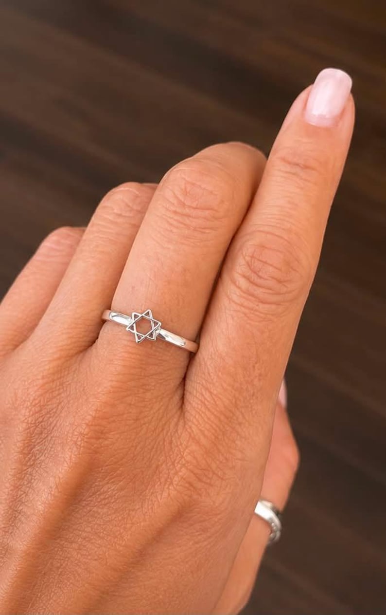 Puede incluir: Un anillo de plata pulida que presenta un peque&ntilde;o s&iacute;mbolo de la Estrella de David calado, centrado en una banda fina y lisa. El metal tiene un acabado met&aacute;lico reflectante y el dise&ntilde;o consta de dos tri&aacute;ngulos entrelazados que forman la estrella.