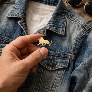 May include: A gold-colored lion pin. The pin is held in front of a blue denim jacket. The lion is in a side profile, with its legs in a walking position. Sunglasses are in the background.