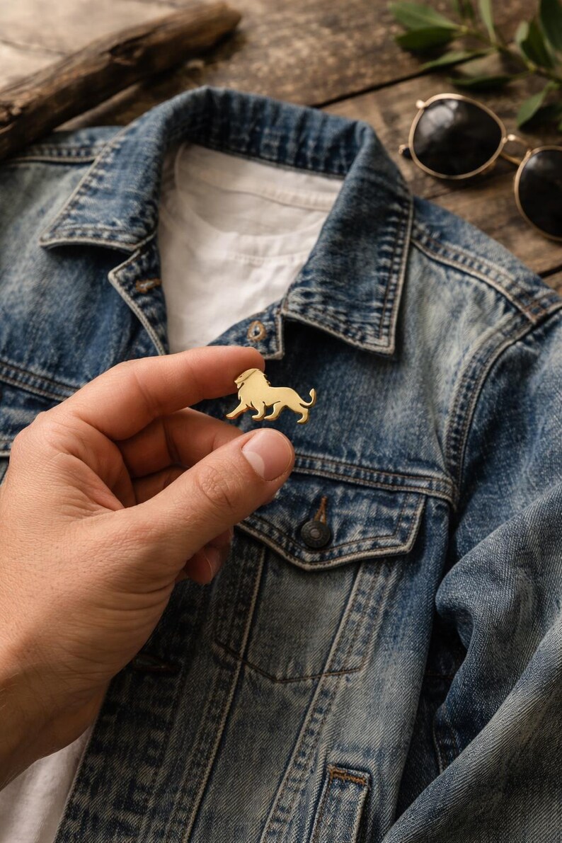 May include: A gold-colored lion pin. The pin is held in front of a blue denim jacket. The lion is in a side profile, with its legs in a walking position. Sunglasses are in the background.