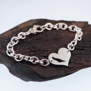 May include: A silver chain link bracelet featuring a polished heart-shaped charm. The charm has a cutout in the centre shaped like the outline of the Sea of Galilee. The bracelet is secured with a lobster claw clasp and has a bright metallic finish.