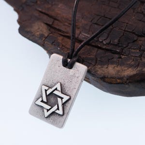May include: A rectangular silver pendant with a Star of David design, suspended from a black leather cord. The pendant has a textured surface and a raised star. The background is a dark, textured wooden surface.