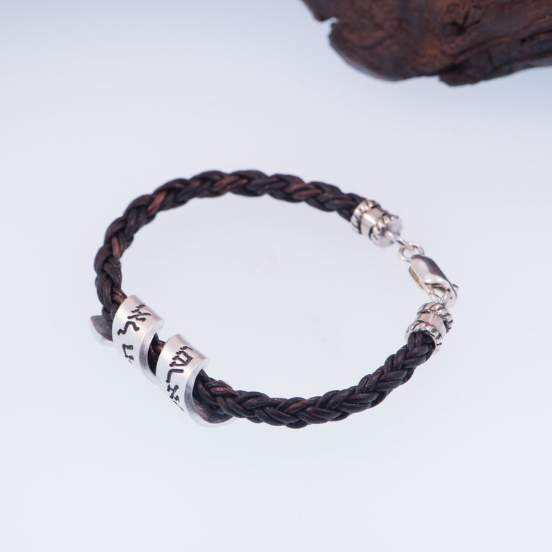 May include: A brown leather braided bracelet with a silver clasp and two silver rings with Hebrew lettering.