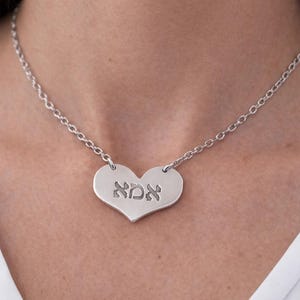 May include: A silver heart-shaped pendant necklace with a chain. The pendant has the Hebrew word "אמא" engraved on it. The necklace is worn around the neck.