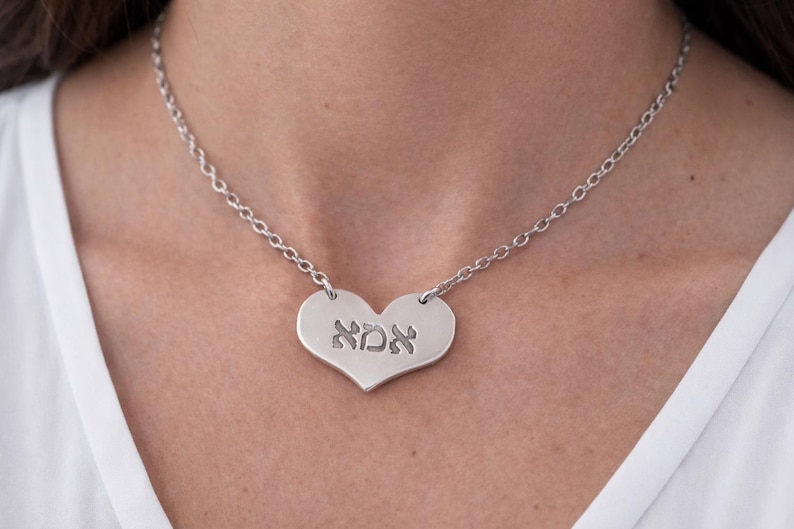 May include: A silver heart-shaped pendant necklace with a chain. The pendant has the Hebrew word "אמא" engraved on it. The necklace is worn around the neck.