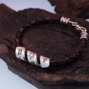 May include: A black leather braided bracelet with silver charms. The charms feature Hebrew letters and a Star of David.
