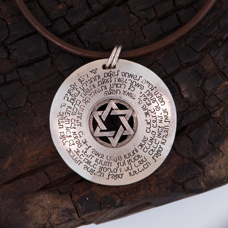 Puede incluir: Collar con colgante de plata. El colgante circular presenta texto hebreo y una Estrella de David. Cuelga de un cord&oacute;n de cuero marr&oacute;n, sobre un fondo oscuro y texturizado.