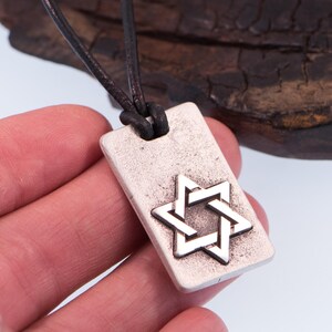 May include: Silver-tone rectangular pendant necklace featuring a Star of David design. The pendant is suspended from a dark brown leather cord. The necklace is a religious symbol and a piece of jewelry.
