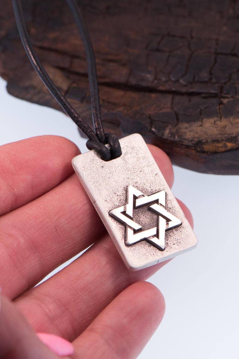 May include: Silver-tone rectangular pendant necklace featuring a Star of David design. The pendant is suspended from a dark brown leather cord. The necklace is a religious symbol and a piece of jewelry.