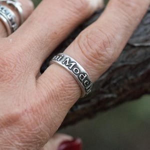 Puede incluir: Un anillo plateado con las palabras "Maui Model" grabadas alrededor de la banda. El anillo se lleva en un dedo, con otros anillos visibles en el fondo. El anillo tiene un estilo vintage.