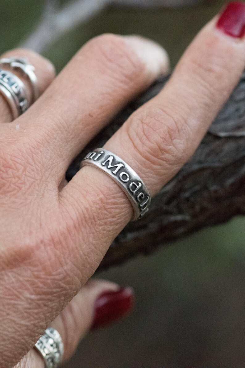Puede incluir: Un anillo plateado con las palabras "Maui Model" grabadas alrededor de la banda. El anillo se lleva en un dedo, con otros anillos visibles en el fondo. El anillo tiene un estilo vintage.