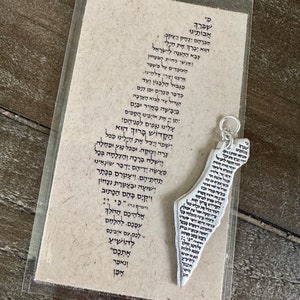 May include: A silver-colored pendant in the shape of a map of Israel, inscribed with Hebrew text, hangs next to a card with more Hebrew text. The card is in a clear plastic sleeve, set against a wooden surface.