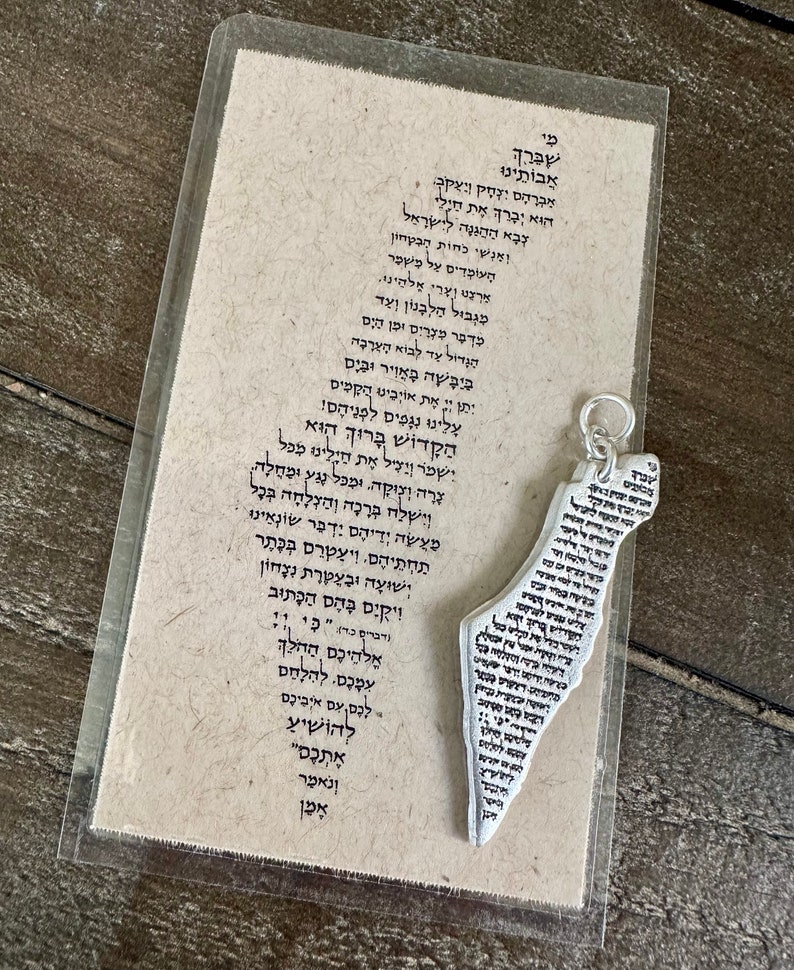 May include: A silver-colored pendant in the shape of a map of Israel, inscribed with Hebrew text, hangs next to a card with more Hebrew text. The card is in a clear plastic sleeve, set against a wooden surface.