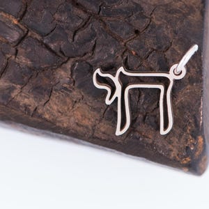 Peut inclure: Un pendentif argent&eacute; en forme du mot h&eacute;breu "Chai" signifiant "vie". Le pendentif est pr&eacute;sent&eacute; sur une surface en bois sombre et textur&eacute;e. Le pendentif a une petite boucle pour une cha&icirc;ne.