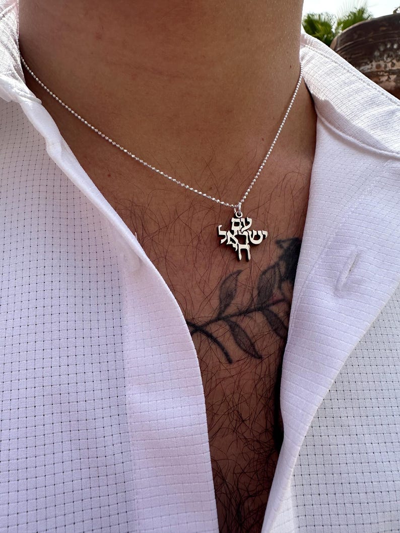 Puede incluir: Un collar de cadena de plata con un colgante con texto en hebreo. El colgante tiene forma de cruz.