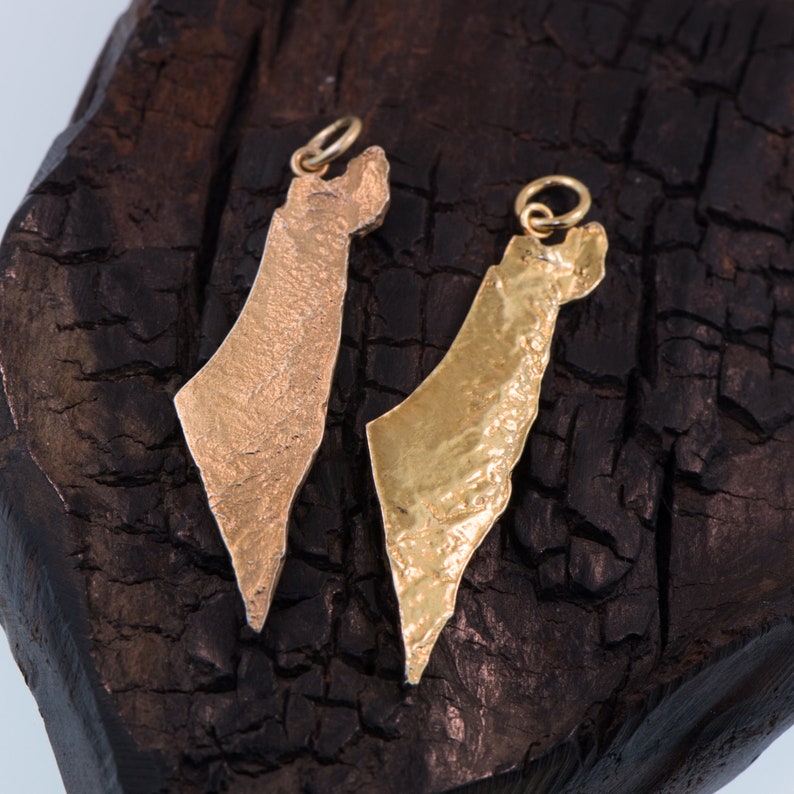 May include: Two metal pendants shaped like the map of Israel, each featuring a textured surface and a small circular jump ring at the top. One pendant has a matte, copper-toned finish, while the other has a bright, polished gold-toned finish.