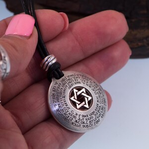Puede incluir: Un collar con colgante plateado con un cord&oacute;n de cuero negro. El colgante redondo presenta una Estrella de David en el centro y texto alrededor del borde. El collar se sostiene en una mano con esmalte de u&ntilde;as rosa.
