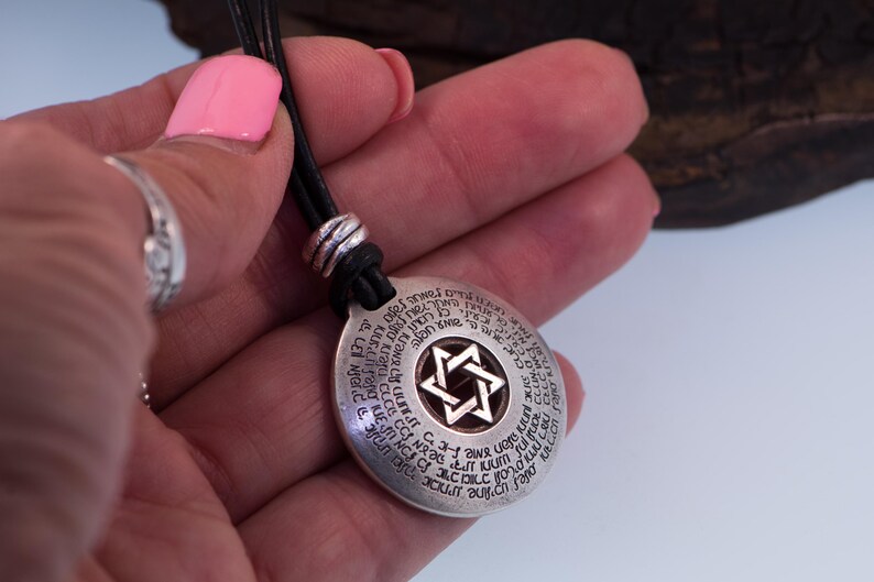 Puede incluir: Un collar con colgante plateado con un cord&oacute;n de cuero negro. El colgante redondo presenta una Estrella de David en el centro y texto alrededor del borde. El collar se sostiene en una mano con esmalte de u&ntilde;as rosa.