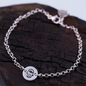 May include: A silver chain bracelet with a hamsa hand charm. The charm is round and has a detailed design of a hand with an eye in the palm.