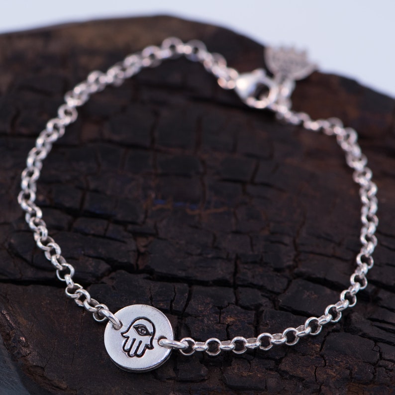 May include: A silver chain bracelet with a hamsa hand charm. The charm is round and has a detailed design of a hand with an eye in the palm.