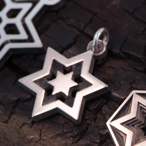 May include: A silver star-shaped pendant with a layered design, featuring a smaller star within. The jewellery is shown against a dark, textured backdrop. The pendant has a loop for a chain.
