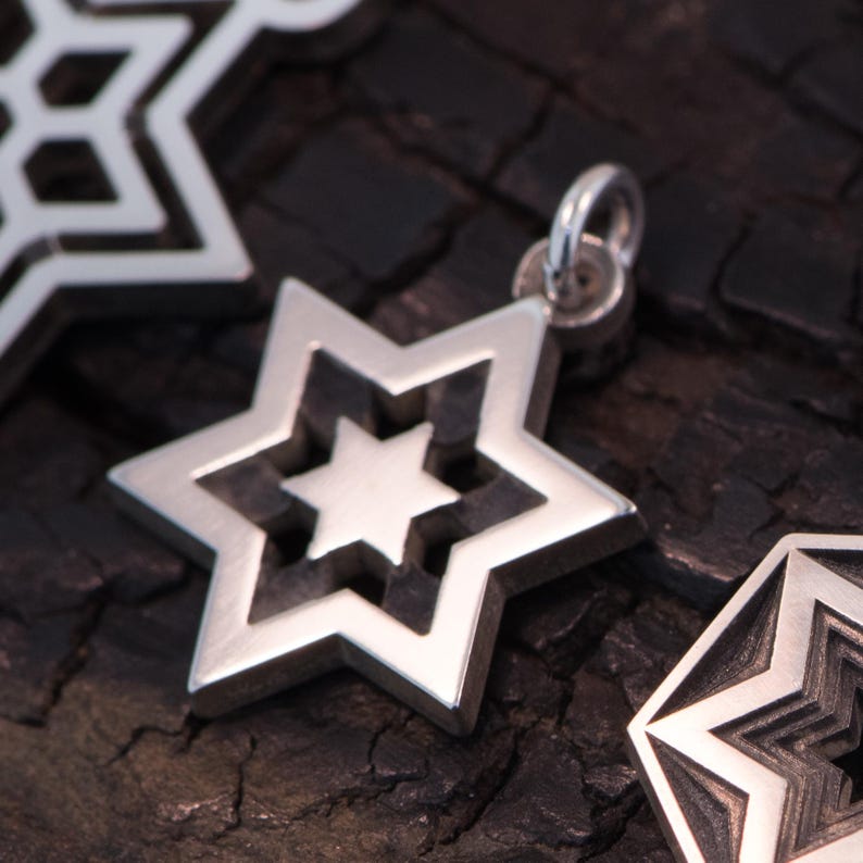 May include: A silver star-shaped pendant with a layered design, featuring a smaller star within. The jewellery is shown against a dark, textured backdrop. The pendant has a loop for a chain.